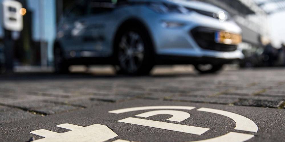 Markering van een parkeerplaats met oplaadsymbool op de grond, met een elektrische auto op de achtergrond.