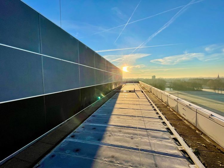 Zonnepanelen hoog op de gevel bij het dak van het provinciehuis, met daarboven een blauwe lucht, de zon en enkele kleine wolken in de verte.