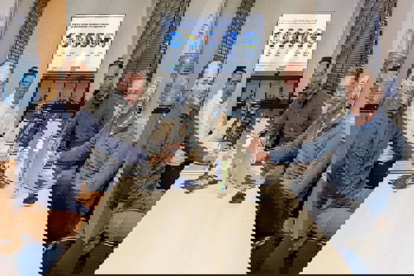 De vier mannen samen rond één lange tafel. In gesprekssetting op de tafel laptops en twee drinkflessen. Achter hen staat een beeldscherm met daarop technische uitleg over de technologie van NEWES in het Engels.