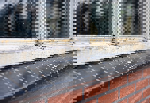 Close-up van een afgebladderd houten raamkozijn dat aan het rotten is en vervangen moet worden, net als de ramen van enkel glas. Onder het kozijn zit een dorpel van zwarte stenen op de muur van rode bakstenen.