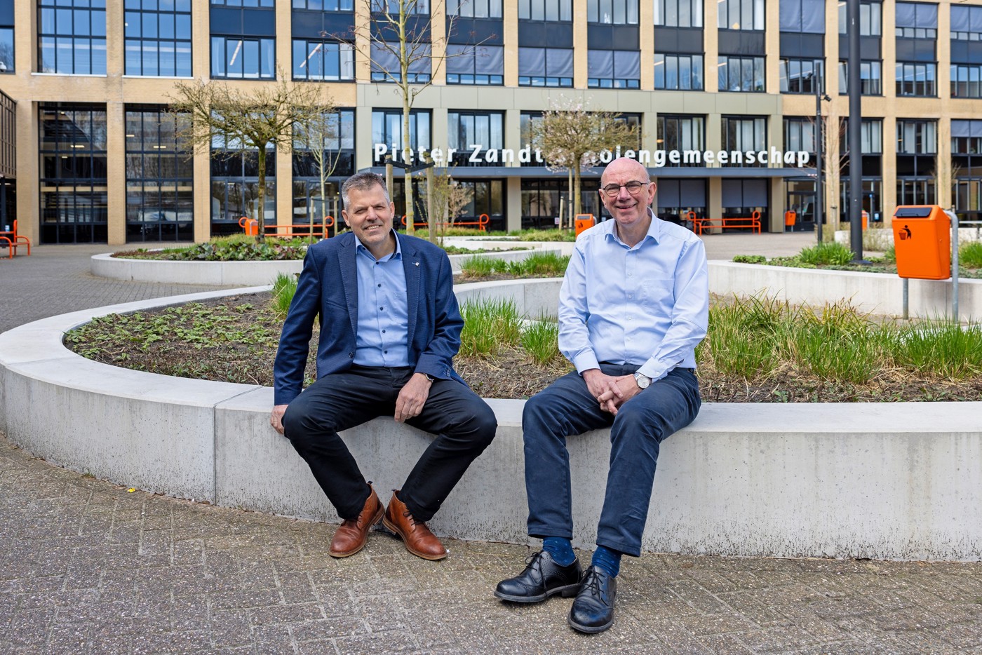 Jelle de Boer en Leen van Wijngaarden zittend op een muurtje op een verder verlaten schoolplein bij een van de schoolgebouwen van het Pieter Zandt. Op de gevel van het gebouw achter hen staat de tekst "Pieter Zandt Scholengemeenschap". Ze zitten op een muurtje van een grote plantenbak met wat kleine groene planten.