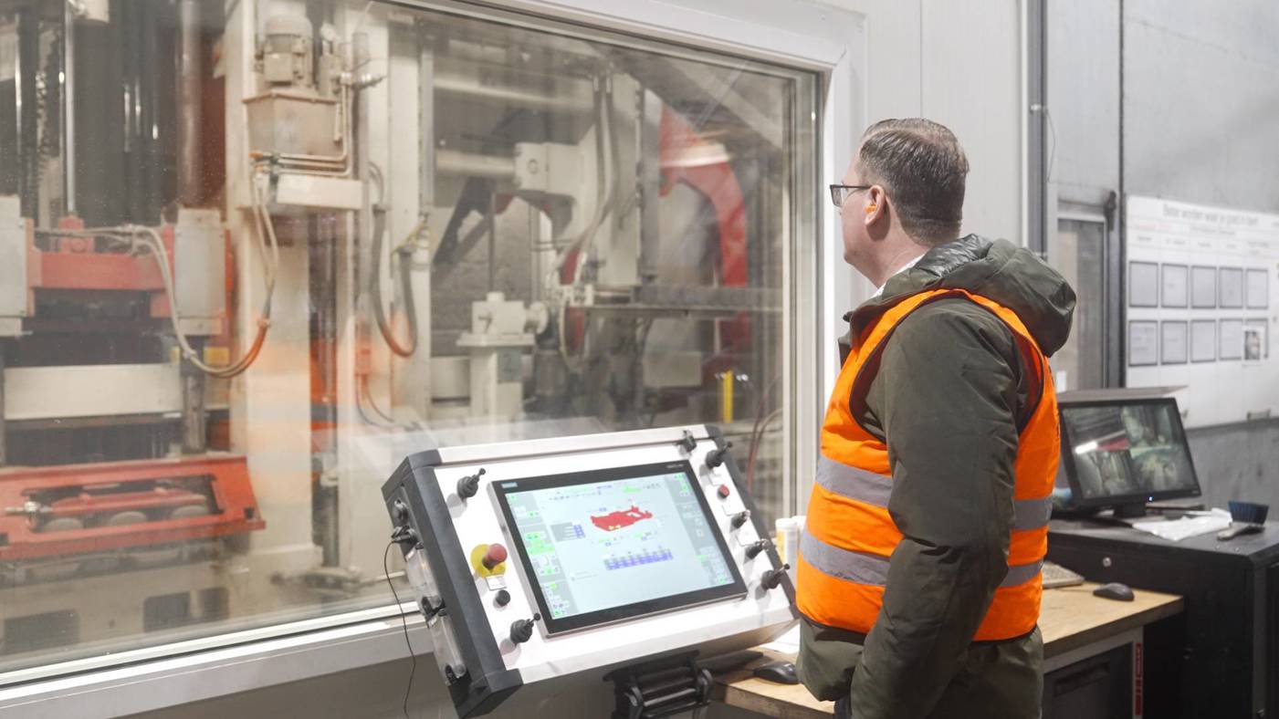 Geert Morssinkhof in een oranje veiligheidsvest kijkt door een raam naar een grote industriële machine en bedient een besturingspaneel.