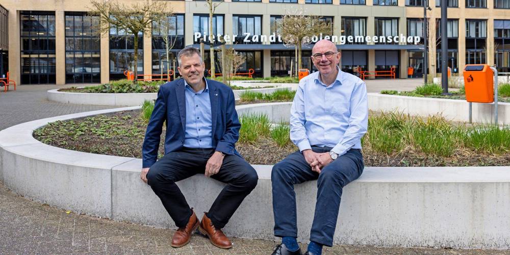 Jelle En Leen zitten op een betonnen rand voor het gebouw van de Pieter Zandt Scholengemeenschap.
