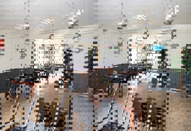 De binnenkant van het verlaten buurthuis met een ouderwets systeemplafond en een oude houten vloer. Er staan houten tafels tegen elkaar aangeschoven met daar omheen plaats voor vier houten stoelen. De meeste stoelen staan nu omgekeerd op de tafels. Aan het plafond hangen zilverkleurige hanglampen.