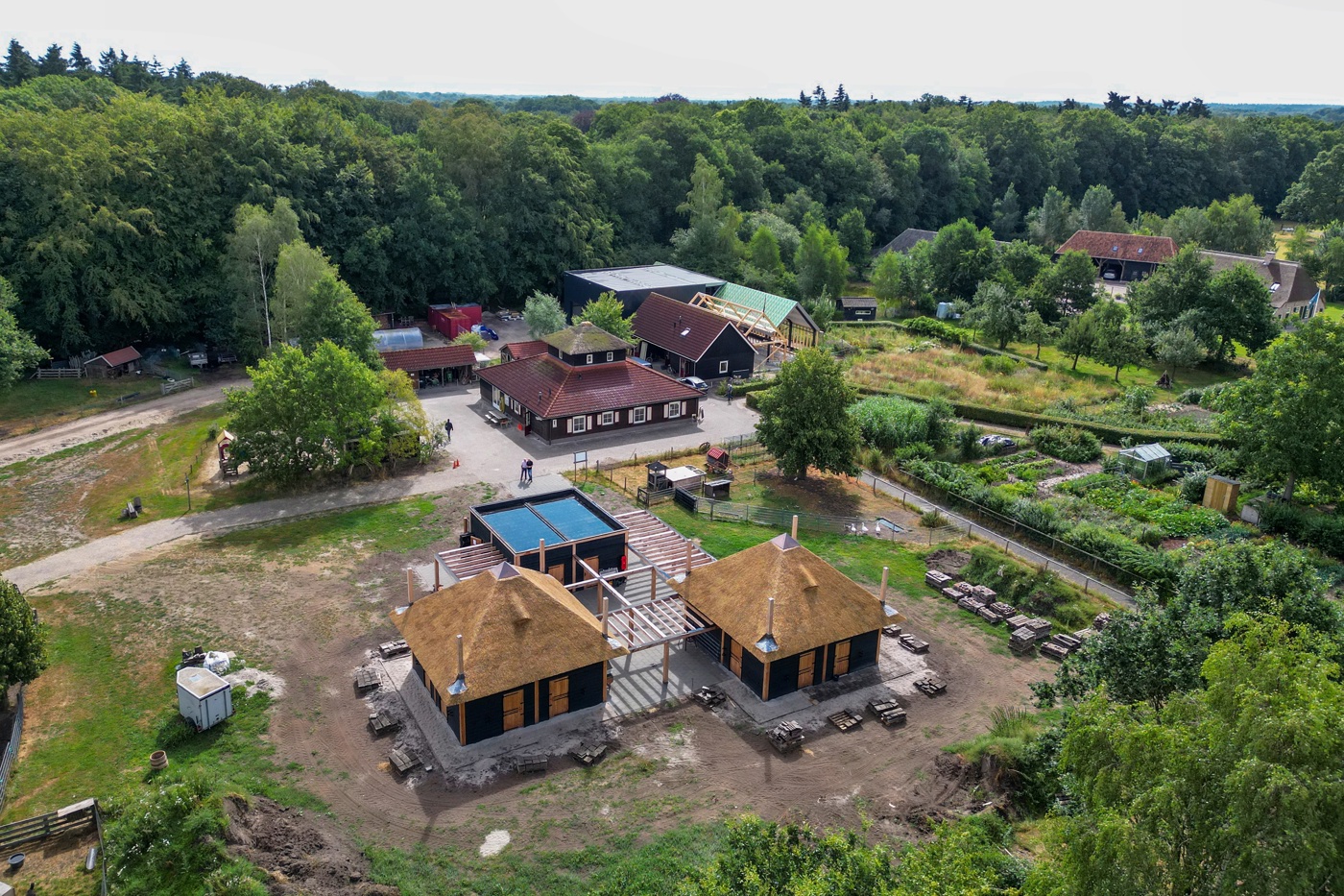 Het erf van de natuurboerderij vanuit de lucht, omgeven door bos. Naast het karakteristieke hoofdgebouw dat van bovenaf vierkant is, op de voorgrond ook de nieuwbouw van drie bijgebouwen die van bovenaf ook vierkant zijn.  