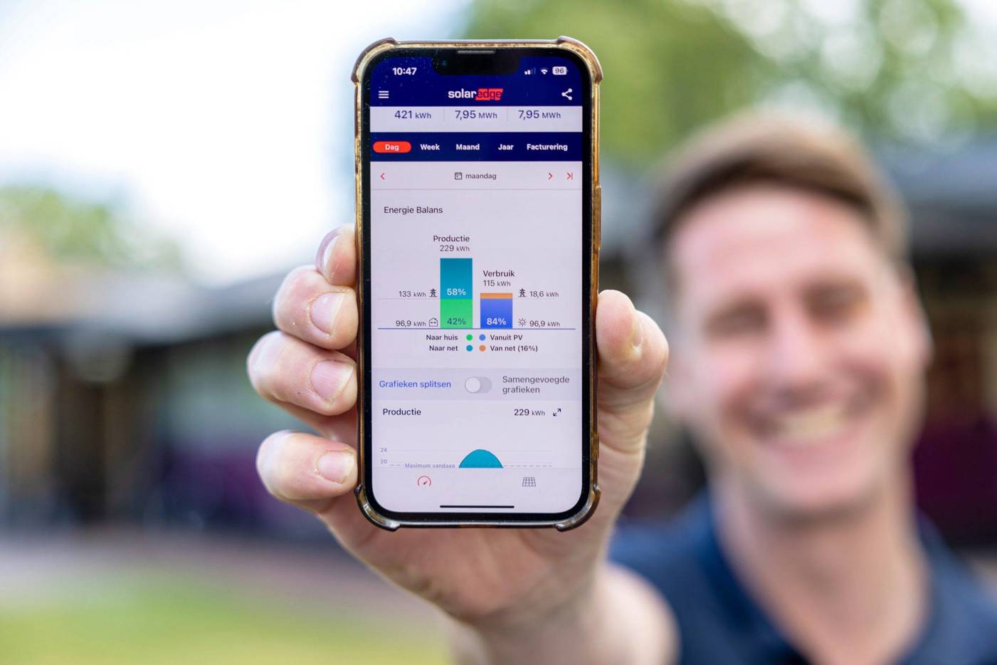 Een hand houdt een smartphone omhoog waarop een energiebeheer‑app wordt weergegeven, buiten in een groene omgeving.