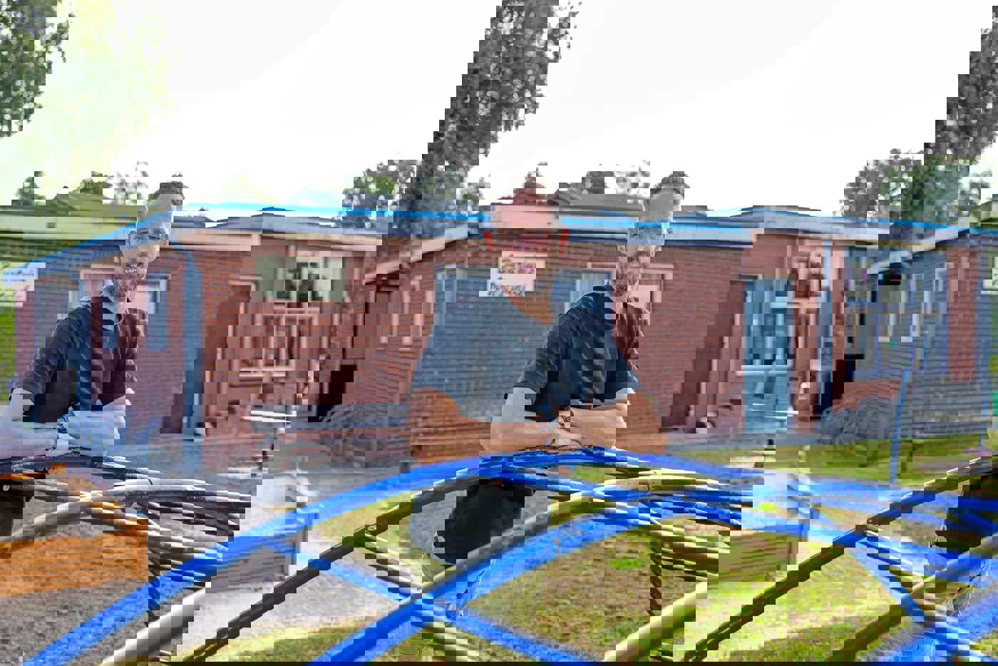 De voorzitter staat buiten op het gras van het speelterrein bij het buurthuis. Hij leunt in zijn T-shirt op een blauw klimtoestel. Achter hem het buurthuis met op de gevel een naambordje met de tekst: "Buurthuis Westerdok." Links achter hem zien we ook een stukje van een houten zandbak.