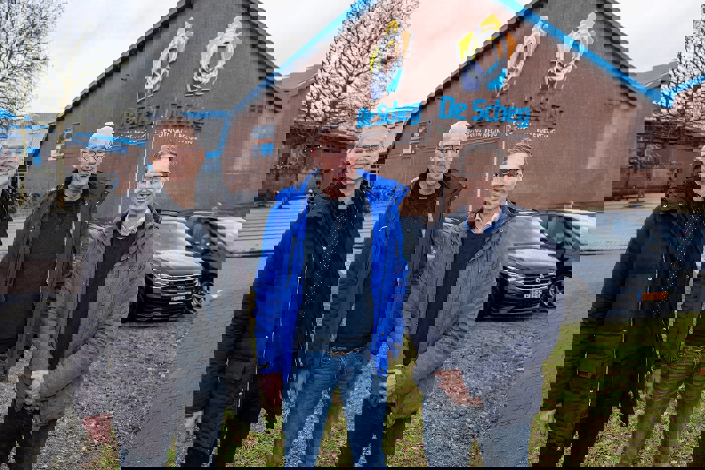 Jaap, Tonnie en Hans op de parkeerplaats voor zwembad De Scheg. Op de gevel van het zwembad staat de naam De Scheg en daaronder de slogan 'Beleving in beweging'. Op de stoep achter de heren staan twee kale bomen.