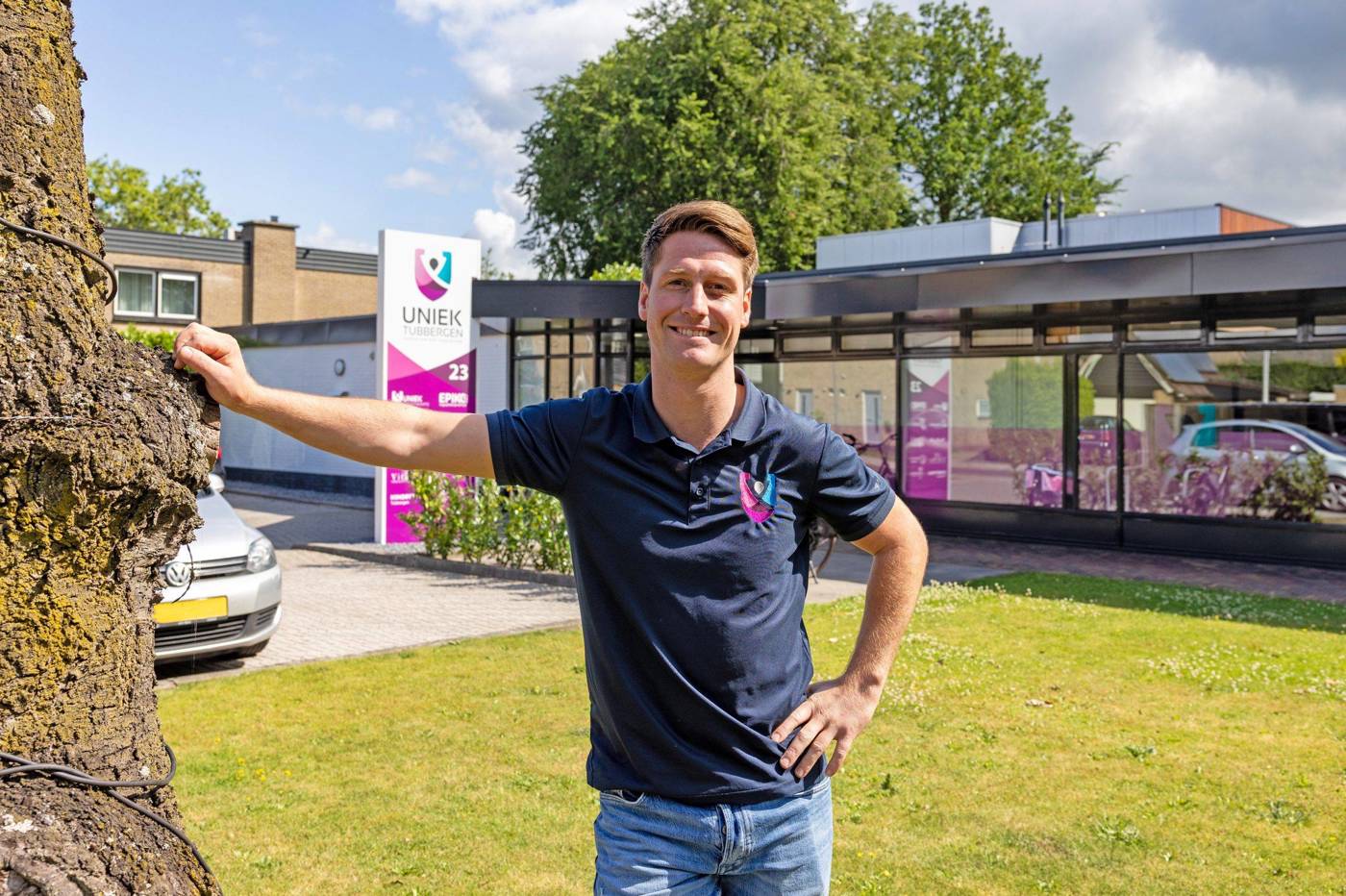 Tim Waaijer in een polo met logo poseert buiten bij een gebouw met een bord waarop “UNIEK Tubbergen” staat.