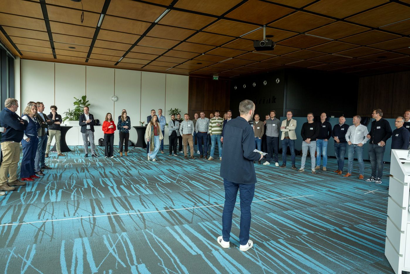 Deelnemers in een zaal van het provinciehuis. Ze staan in een kring rond een spreker te luisteren. In de afbeelding van de  blauwgroene vloerbedekking is structuur van riet te zien.
