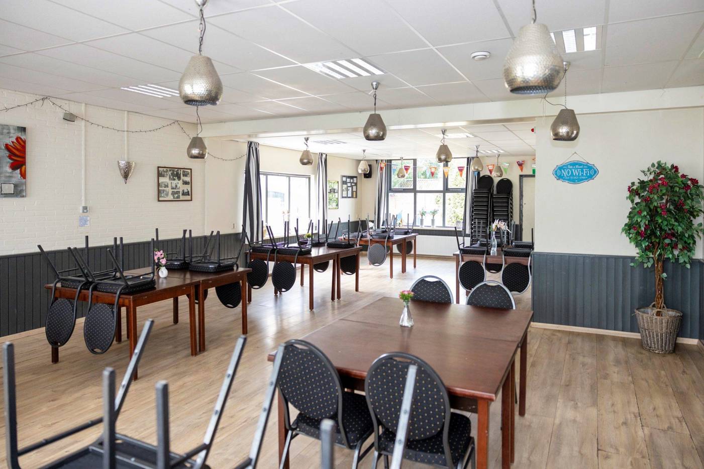 Een grote zaal in een buurthuis met houten tafels, stoelen die ondersteboven op de tafels staan, hanglampen en grote ramen die daglicht binnenlaten.