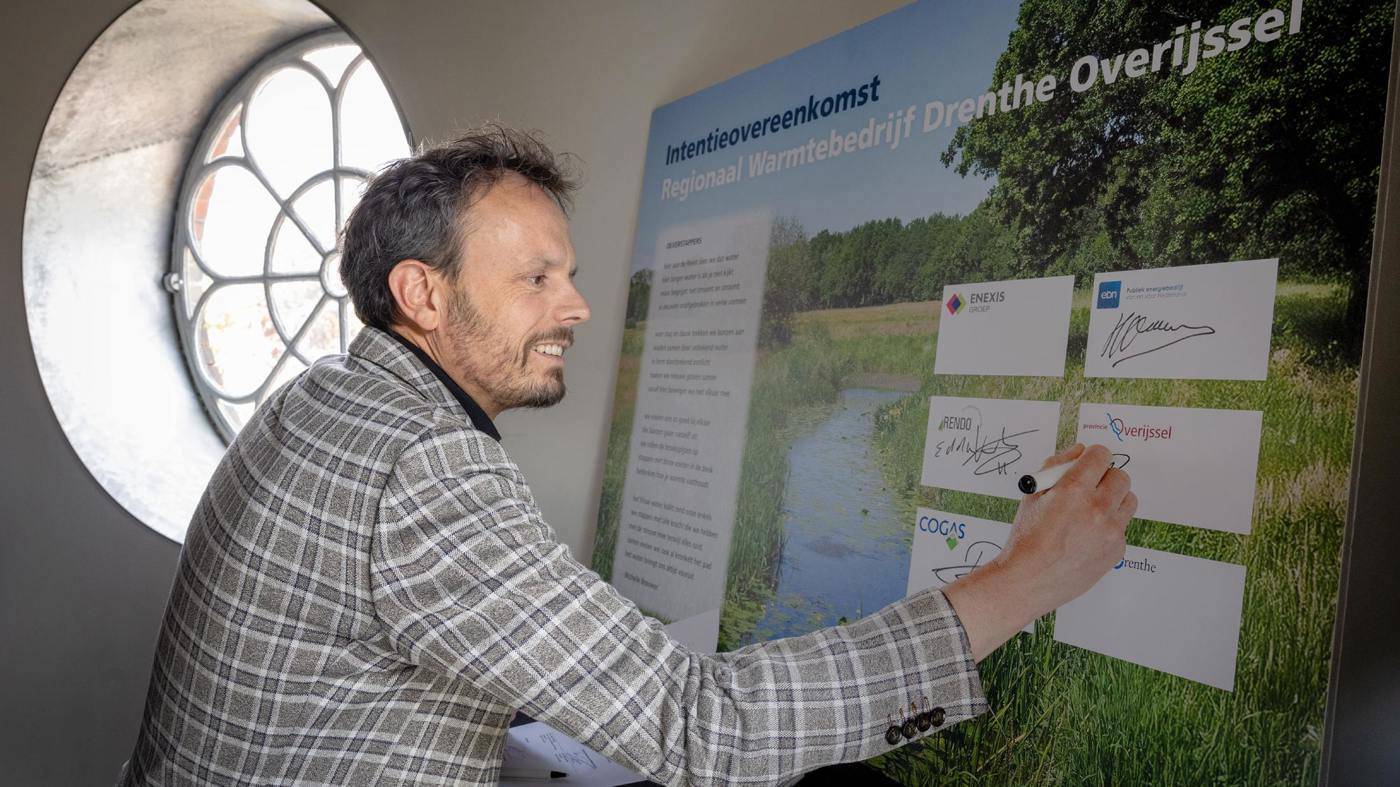 Gedeputeerde Tijs de Bree ondertekent een groot bord met de titel “Intentieovereenkomst Regionaal Warmtebedrijf Drenthe‑Overijssel”, met een landschapfoto en meerdere logo’s en handtekeningen.