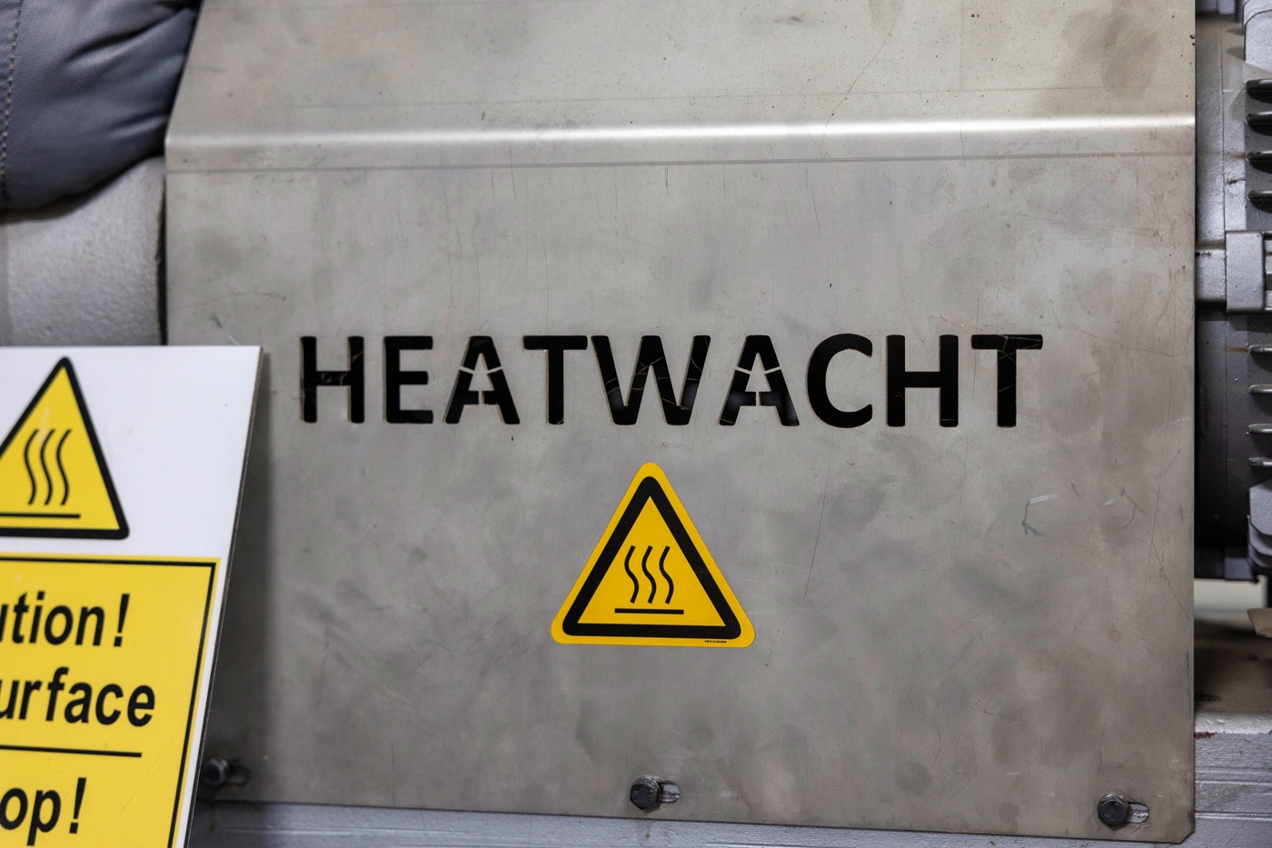 Close-up van een onderdeel van de installatie met daarop de bedrijfsnaam Heatwacht en een gele waarschuwingssticker dat het onderdeel warm kan zijn.