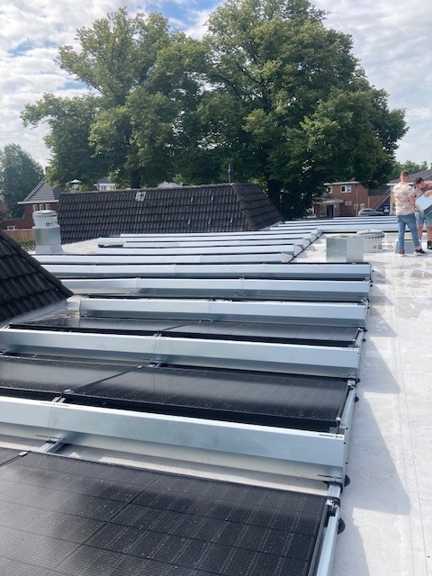 Zij-aanzicht van de zonnepanelen op het nieuwe dak van het pand. Op de achtergrond zijn twee mensen aan het werk op het dak van 't Brookshoes.Nog verder weg zijn twee bomen vol groene bladeren te zien en de daken van omliggende gebouwen.