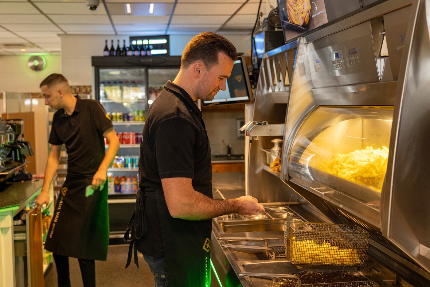 Mark vanaf de zijkant gezien terwijl patat bakt bij de frituuroven van het restaurant. Achter hem ook een andere horecamedewerker en een koelkast met frisdrank.