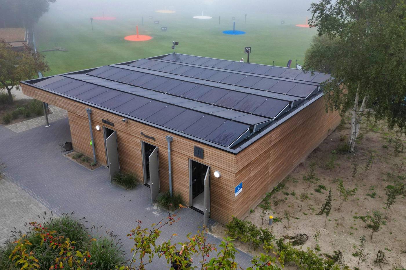 Houten gebouw met meerdere deuren waarvan enkele openstaan, met een dak volledig bedekt met zonnepanelen en mistig terrein op de achtergrond.