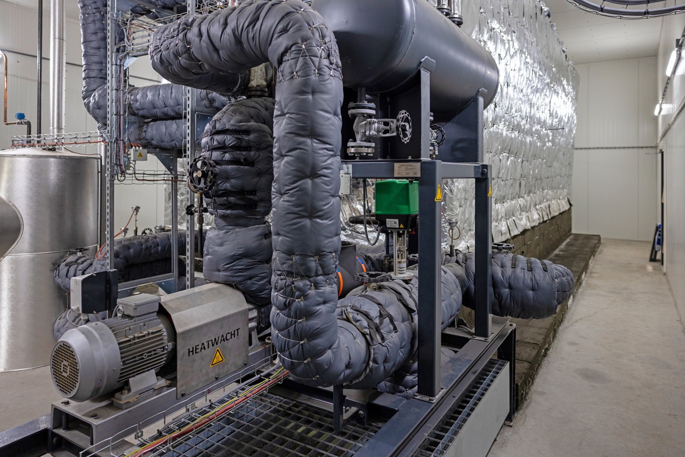Industriële installatie met diverse buizen, koppelingskranen en een hoge vloeistoftank. De warmtebuizen zijn dik ingepakt voor goede warmteisolatie. 