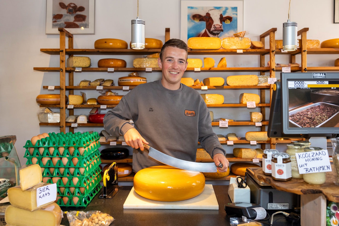 Siebe glimlachend achter de toonbank van de winkel met achter hem een rek met verschillende soorten kazen. Voor hem op de toonbank ligt een grote ronde kaas waar hij het speciale kaasmes in zet. Op de toonbank staan ook dozen met eieren, bierkaas en lokale koolzaadhoning.