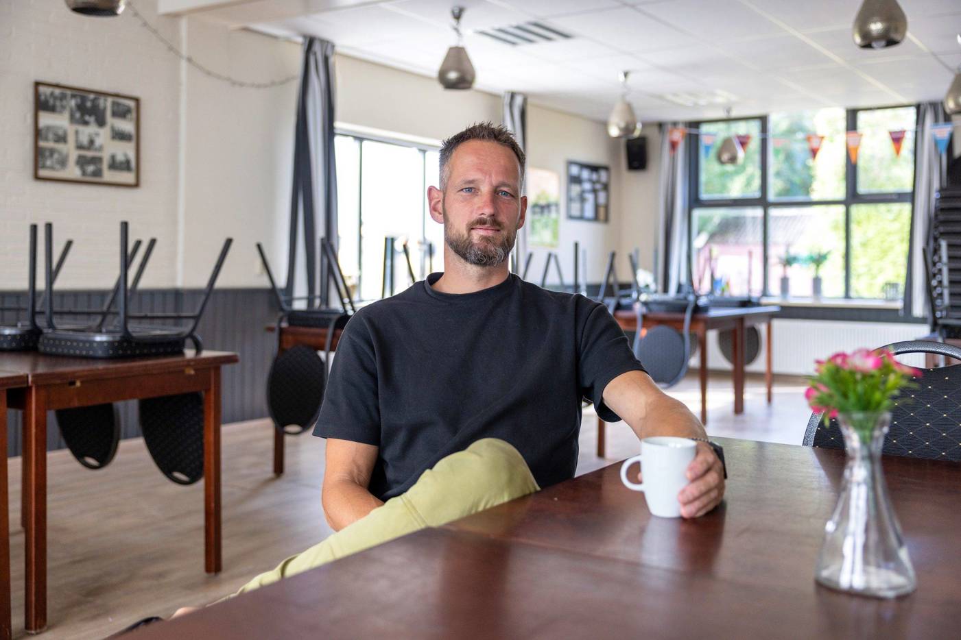 Rik Voerknecht zit ontspannen aan een tafel in een buurthuis en houdt een witte mok vast, terwijl stoelen ondersteboven op tafels staan in de achtergrond.