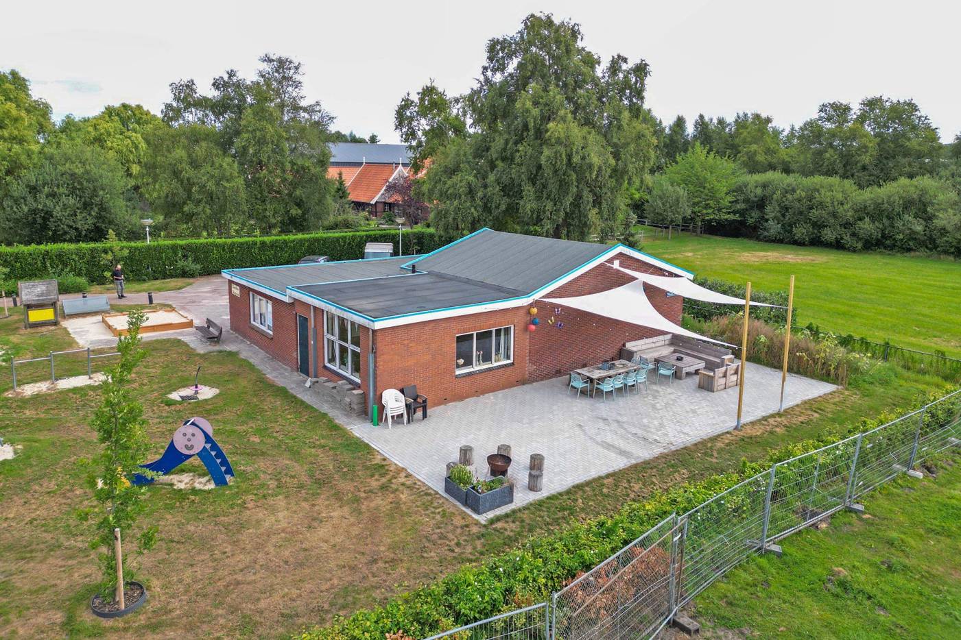 Luchtfoto van een buurthuis met terras, zitplekken, speeltoestellen en een groot groen veld eromheen.